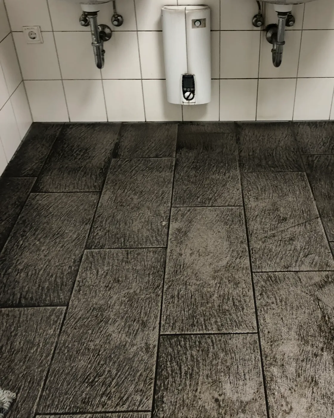 Dunkler, vergrauter Fliesenboden in einem Sanitärraum vor der Grundreinigung
