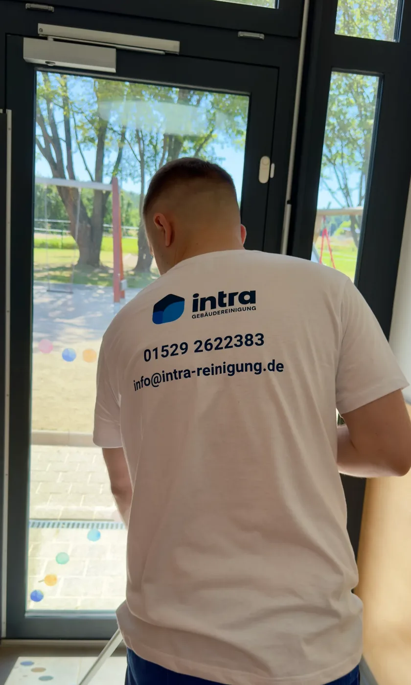 Mitarbeiter von intra reinigt die Glastür einer Kita, auf dem Shirt stehen Telefonnummer und Mailadresse